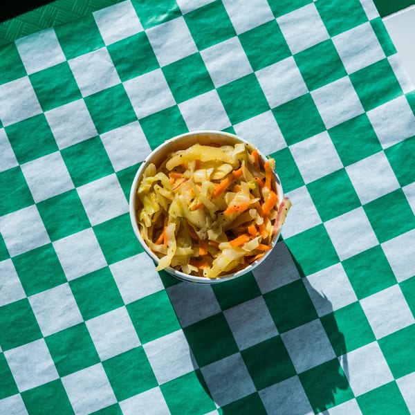 A white paper cup filled with coleslaw, featuring shredded cabbage and carrots, on a green and white checkered paper.