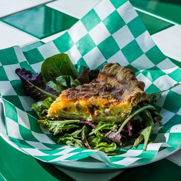 A slice of quiche with a side salad in a green and white checkered paper-lined metal dish.