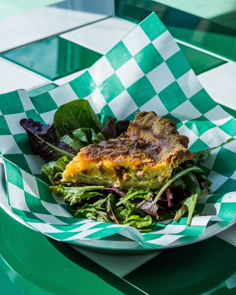 A slice of quiche with a side salad, served on green and white checkered paper.