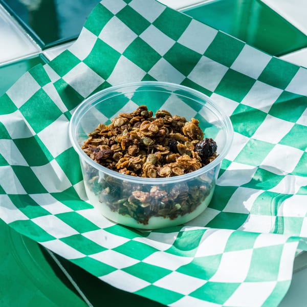 Granola with yogurt in a clear plastic cup, on green and white checkered paper.