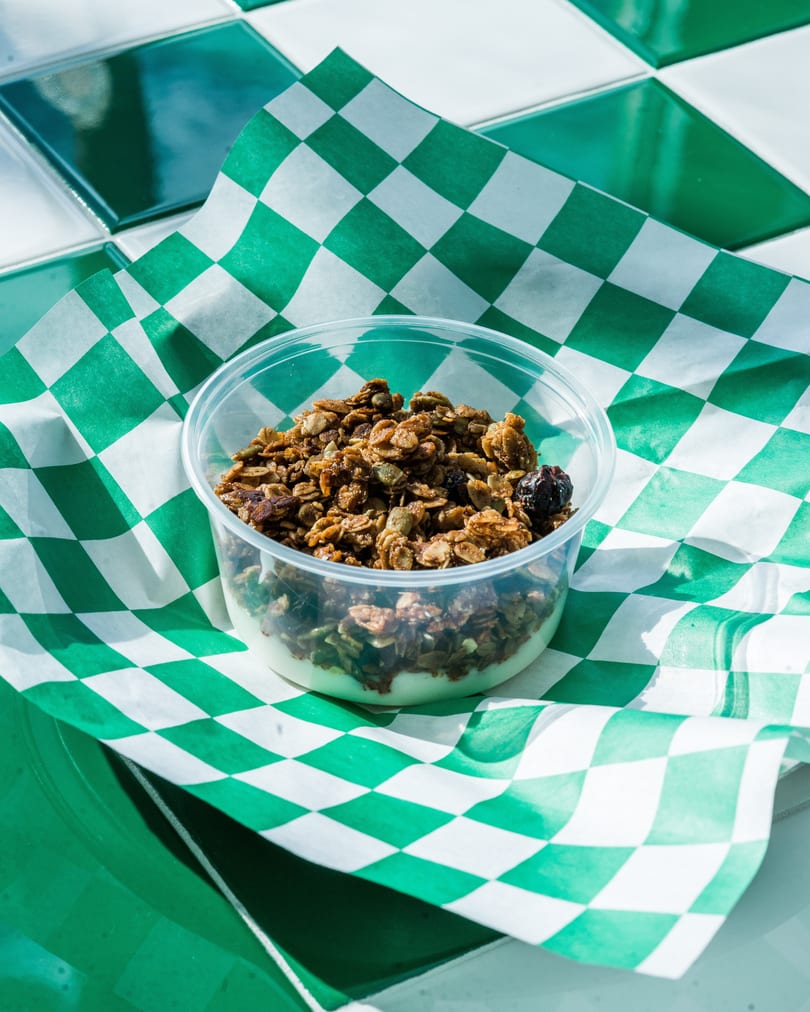 Granola with yogurt in a clear plastic cup, on green and white checkered paper.