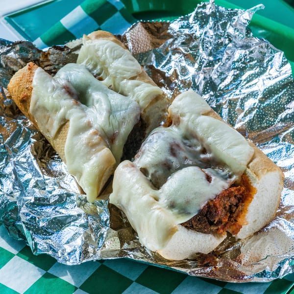 Two cheesy meatball subs on a green checkered paper liner and aluminum foil.