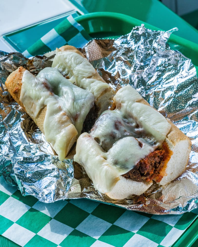 Two cheesy meatball subs on a green checkered paper liner and aluminum foil.